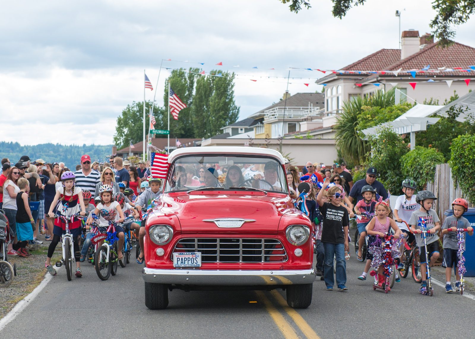 The Fourth of July parade on Maplewild Ave SW at Three Tree Point — a vintage red Chevy pickup leading kids on flag-decorated bikes and scooters past crowds lining the street.