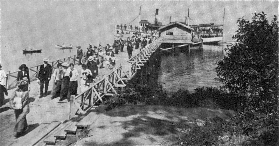 Summer visitors walking the pier at the Three Tree Point landing in the early 1900s, with a Mosquito Fleet steamer moored alongside.